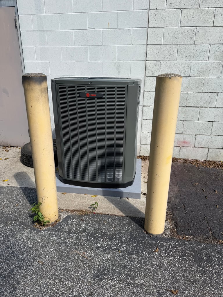 HVAC air conditioning unit mounted against a white concrete block wall, flanked by two yellow protective bollard posts on asphalt ground.