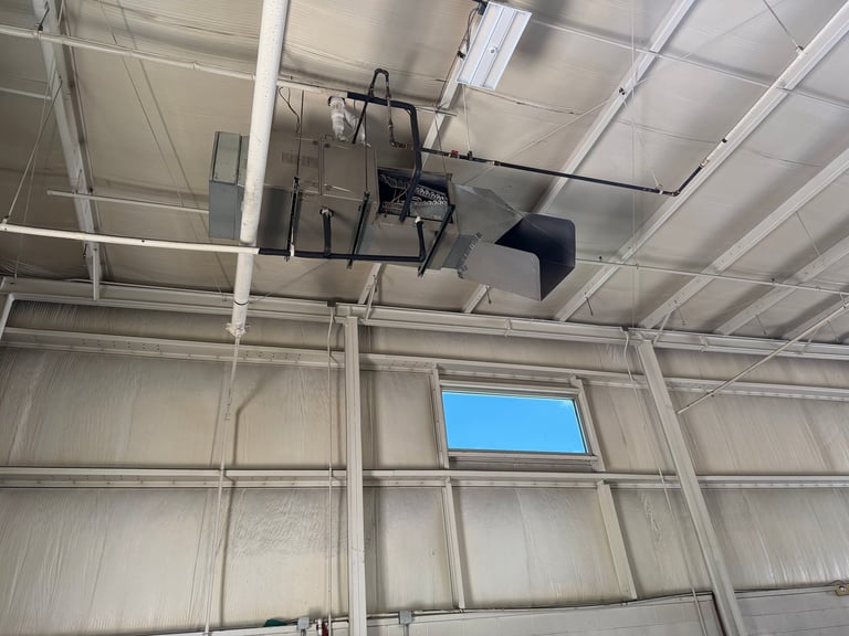Industrial metal garage or warehouse interior with corrugated walls, exposed ceiling beams, and mounted mechanical equipment including a pulley system and ventilation ductwork. Blue sky visible through a window opening.
