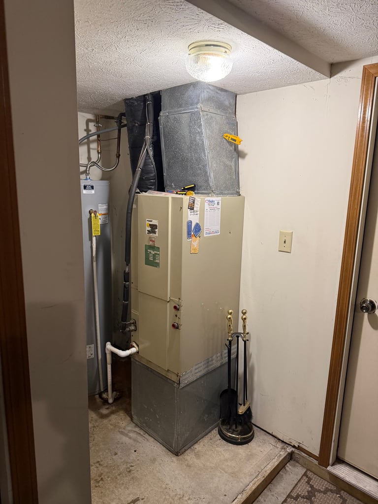 Utility closet with a large beige water heater, gray metal ductwork, pipes, and a plunger against white walls