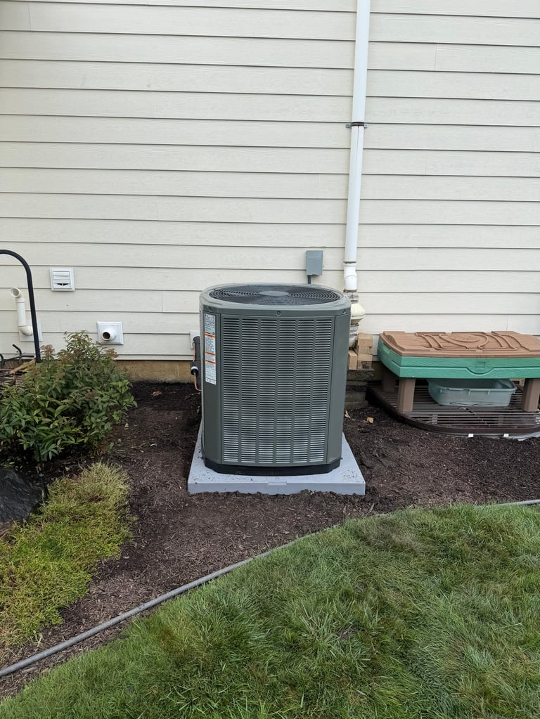 Gray air conditioning unit mounted on concrete pad against white house siding with landscaping
