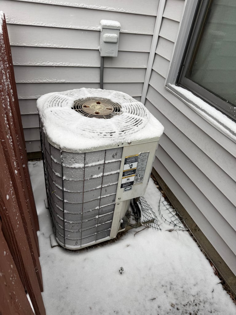 Outdoor air conditioning unit covered in snow and frost next to a residential house wall and window