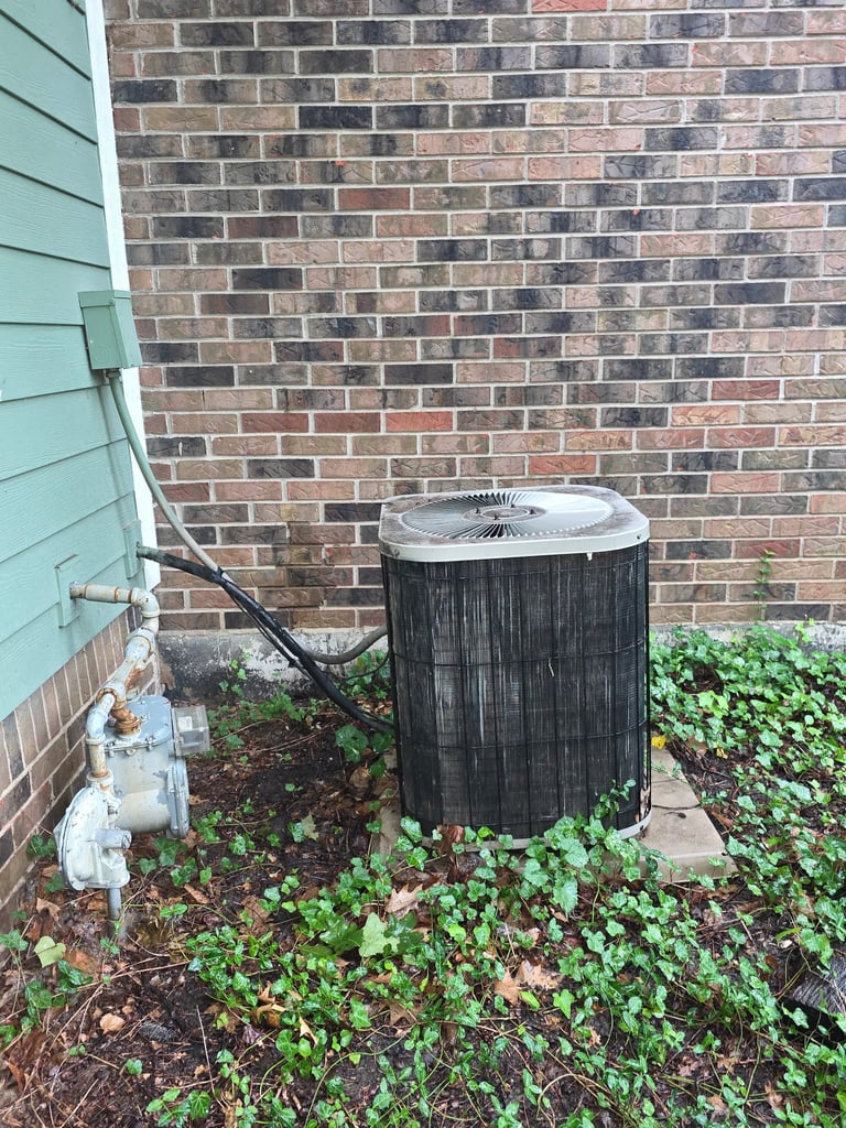 Outdoor air conditioning unit with weathered black cylindrical casing against brick wall, surrounded by green ivy and vines