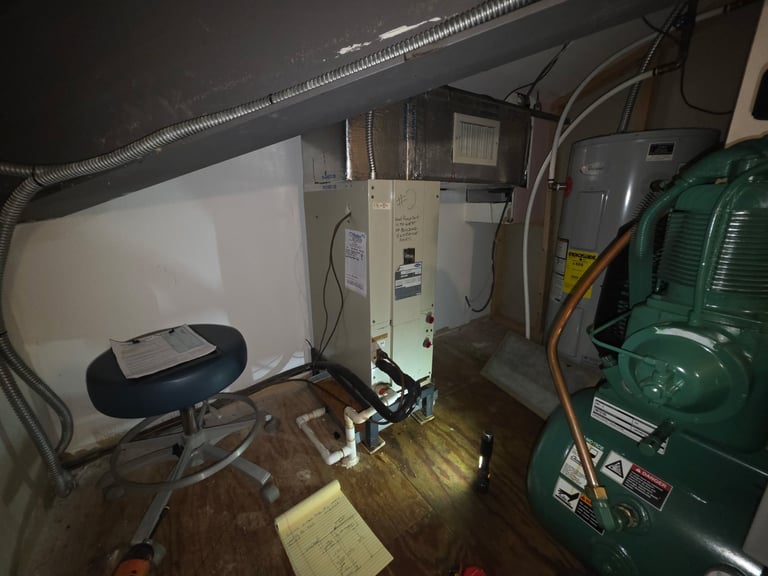 Cluttered mechanical room with HVAC equipment, green compressor, wiring, and various utility systems on wooden flooring
