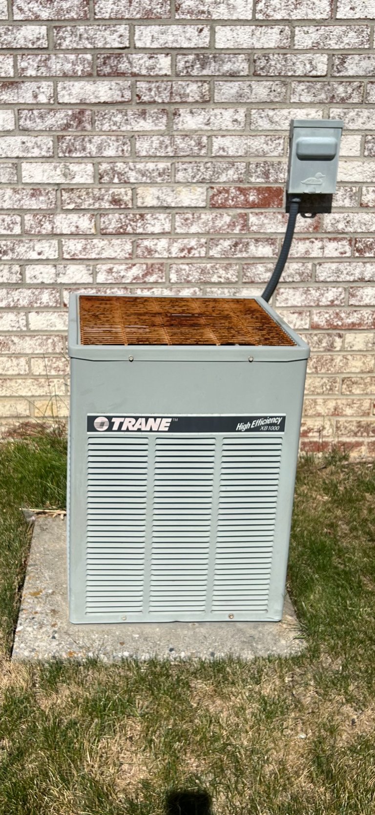 Trane high efficiency air conditioning unit against a brick wall with outdoor disconnect box mounted above it