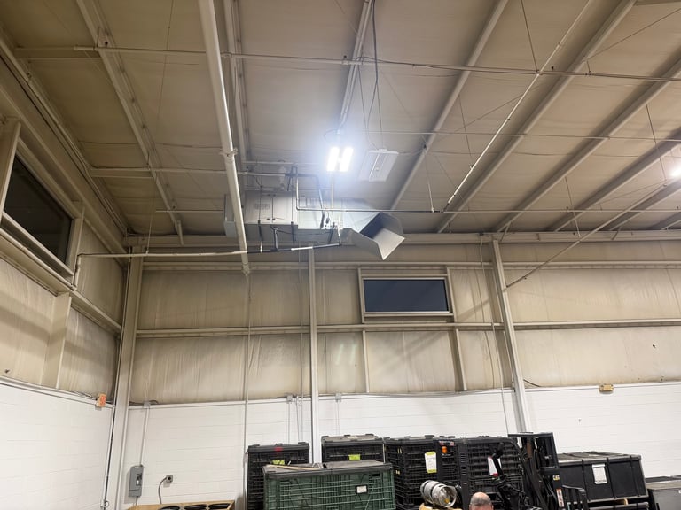 Industrial warehouse interior with metal beam framework, exposed ceiling, fluorescent lights, and stacked equipment or machinery on the floor