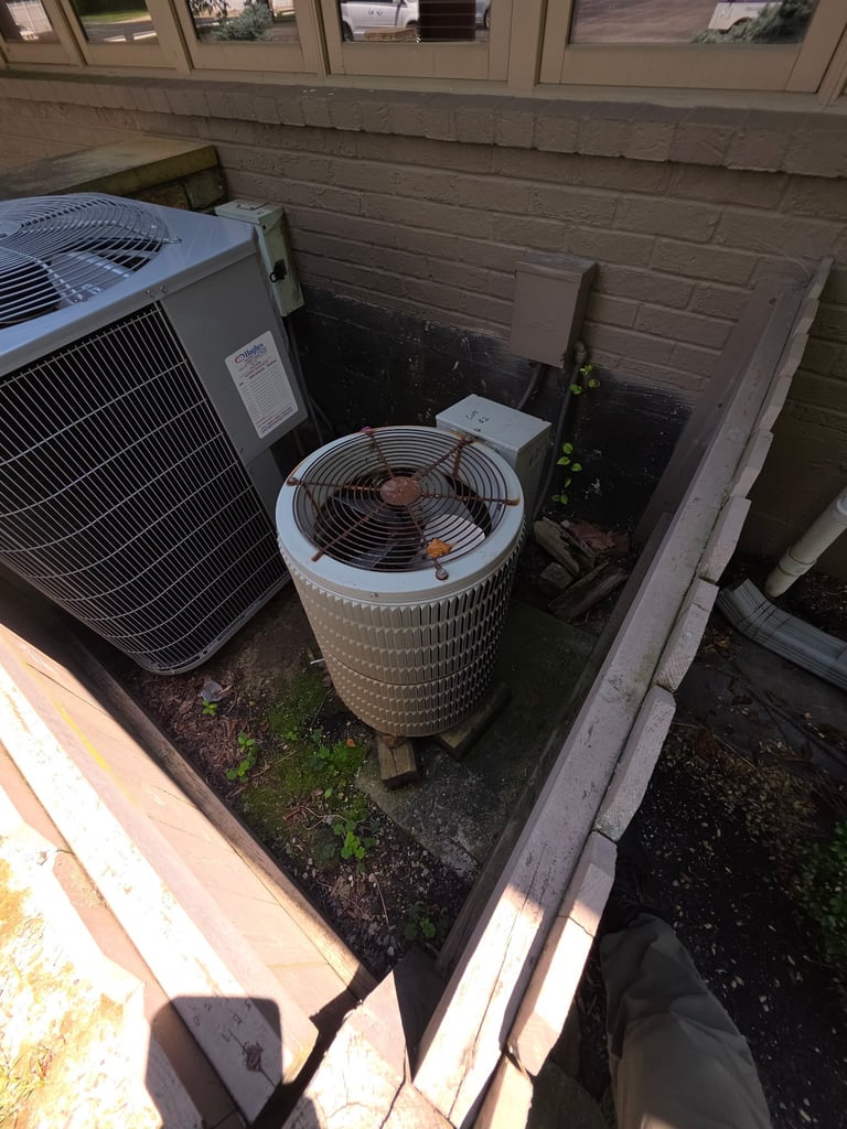 Two outdoor HVAC units in a concrete enclosure with a metal fence, showing an air conditioner compressor and condenser unit next to a brick wall