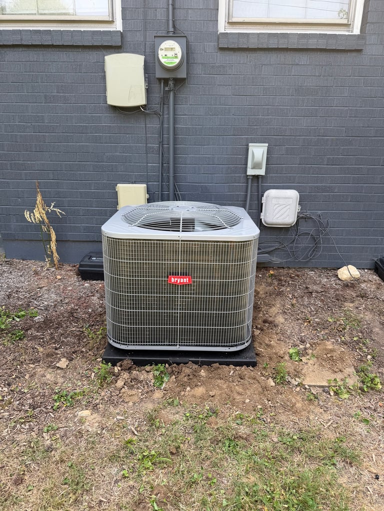 Outdoor HVAC air conditioning unit mounted against a gray shingle exterior wall with electrical components and meter visible above