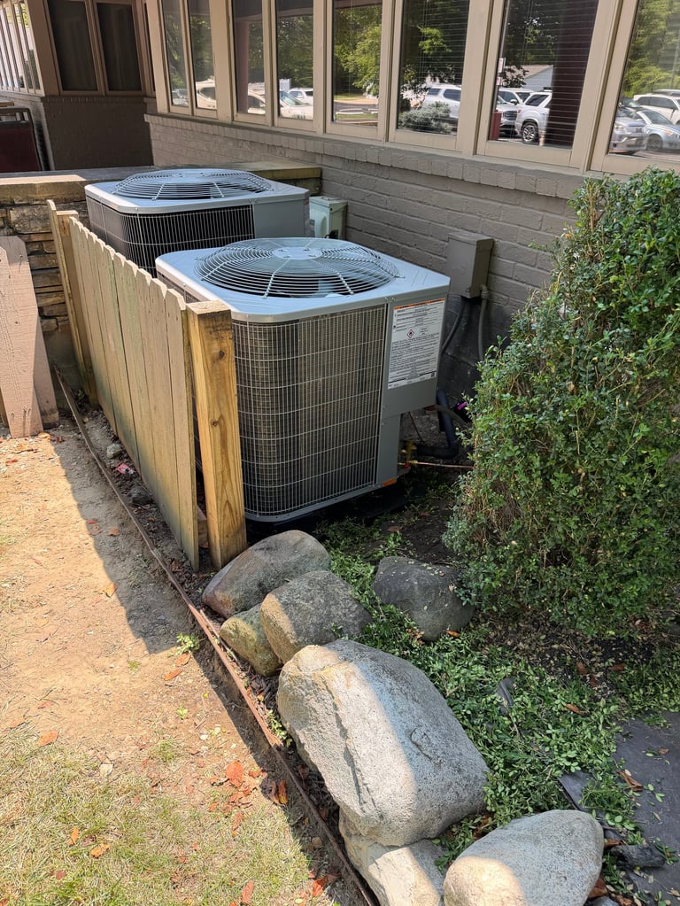 Two outdoor HVAC air conditioning units with metal casing enclosed by wooden screening fence, beside green shrub and stone border