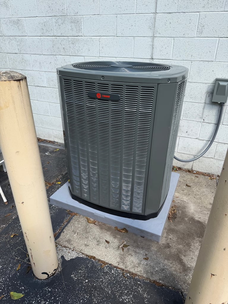 Outdoor HVAC air conditioning unit mounted on concrete pad against white brick wall with electrical conduit and pipes